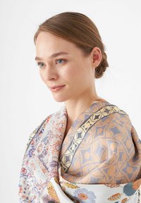 Écharpe en soie ornée de motifs floraux et géométriques complexes dans des tons pastel de bleu, orange et beige, drapée élégamment autour des épaules du mannequin.