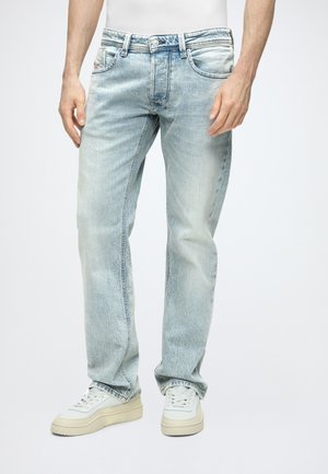 Homme portant un jean droit clair et des baskets blanches, debout devant un fond clair uni.