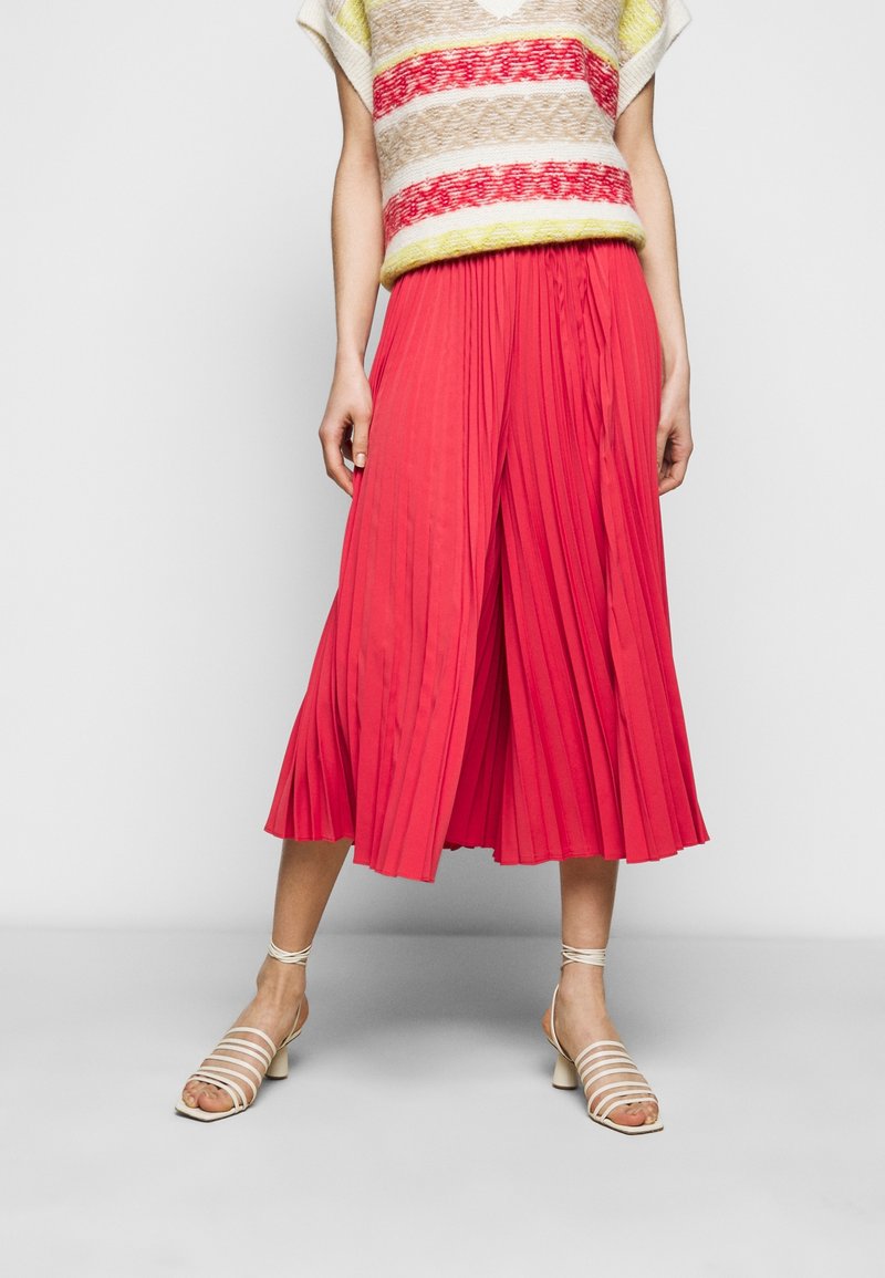 Pleated midi-hame kirkkaassa korallinvärissä, valmistettu kevyestä kankaasta. Yhdistetty neulepuseron kanssa, jossa on vaakaraitoja beigeissä, punaisissa ja keltaisissa väreissä.