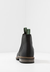 Bota Chelsea de couro preta com painéis laterais elásticos e etiquetas em verde. Apresenta um salto baixo com acabamento de madeira e costura contrastante.