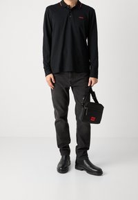 Schwarzes Langarm-Polo mit rotem Kragenrand, schwarze Jeans und schwarze Stiefeletten. Schwarze Umhängetasche mit rotem Logo-Detail.