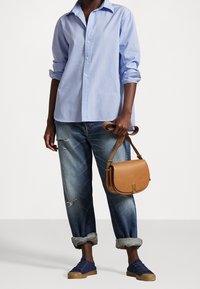 Persona che indossa una camicia azzurro chiaro con bottoni, jeans strappati arrotolati, sneakers blu navy, mentre tiene una borsa a tracolla in pelle beige per la tracolla.