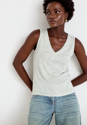 Donna con capelli corti e ricci che indossa orecchini a cerchio dorati, una canottiera grigio chiaro con scollo a V e jeans blu chiaro, in piedi con le mani dietro la schiena.