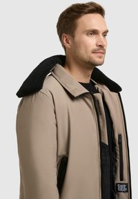 Beige isolert jakke med en svart fleeceforet krage, glidelåslommer og en glatt, elegant tekstur. Har merke på forsiden.