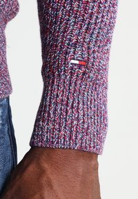 Manche de pull en tricot avec des nuances variables de violet et une texture côtelée, ornée d'un petit écusson logo rouge, blanc et bleu près du poignet.