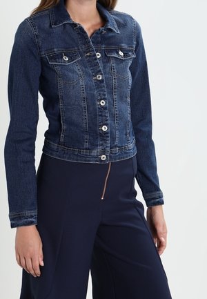 Mujer que lleva una chaqueta vaquera ceñida de color azul oscuro con botones blancos y pantalones azul marino con cremallera frontal visible.