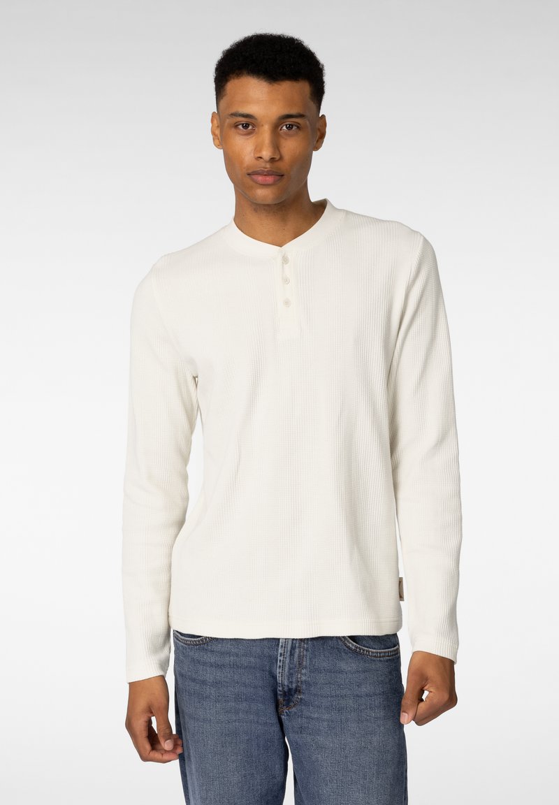 Hvit langermet henley-skjorte laget av teksturert stoff, med tre knapper ved kragen, kombinert med blå denimjeans.