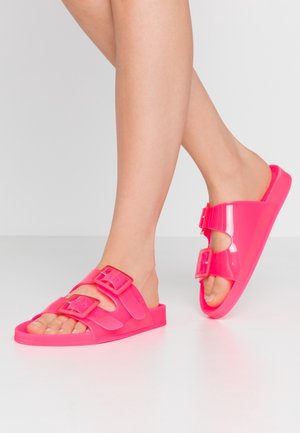 Voeten met felroze open-toe sandalen met twee verstelbare gespsluitingen, staand op een lichtgrijze vloer.