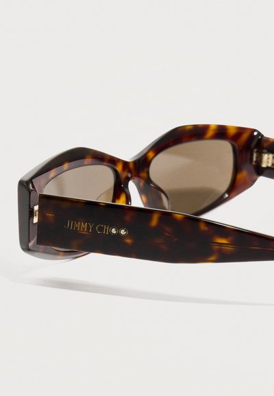 Καφέ γυαλιά ηλίου σε σχήμα γάτας με καφέ φακούς, διακοσμητικά πλαίσια και χρυσαφένια γράμματα "JIMMY CHOO" με κρυστάλλινες λεπτομέρειες.