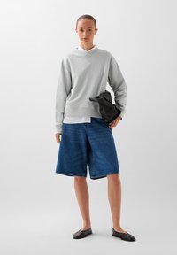 Lichtgrijze sweatshirt met geribde zoom, losse denim shorts in donkerblauw en zwarte platte schoenen. Model houdt een zachte zwarte tas vast.