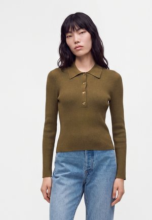 Kvinde med langt sort hår iført en olivengrøn ribbet langærmet poloshirt og lyseblå jeans, stående mod en ensfarvet hvid baggrund.