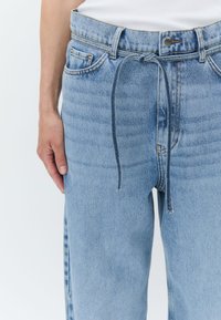 Jean en denim bleu clair avec une coupe décontractée, présentant une fermeture à cordon, des poches avant et des détails de texture verticale subtils.