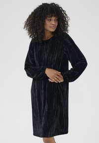 Rochie din catifea neagră, cu mâneci lungi, decolteu rotund și o croială lejeră. Suprafața are un model texturat, cu dungi.