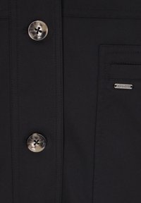 Schwarzer Stoff mit glatter Textur, versehen mit einem großen silbernen Knopf und einem kleinen Markenetikett. Enthält eine seitliche Taschen-Detail.