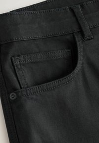 Gros plan sur un jean en denim noir montrant la poche avant, la boucle de ceinture et les coutures détaillées.