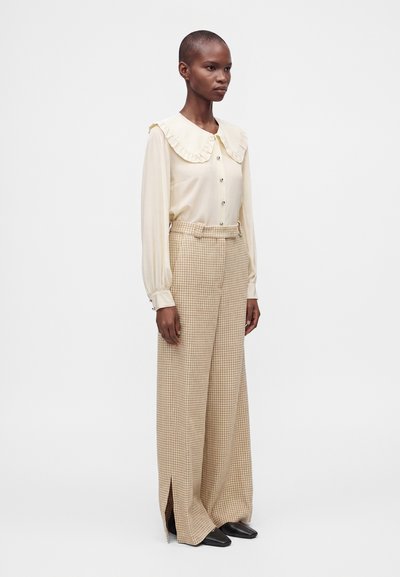 Femme portant un chemisier crème avec un col à volants et des poignets boutonnés, associé à un pantalon large à carreaux beige avec des fentes à l'avant et des chaussures noires.