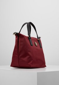 Borsa a mano in nylon rosso con manici in pelle nera; presenta un emblema rotondo dorato e una forma strutturata con texture liscia.