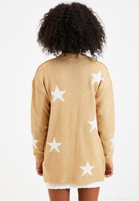 Virkat kofta i beige med vita stjärnmönster, långa ärmar och en bekväm passform. Mjuk textur med en längre bakre fåll.