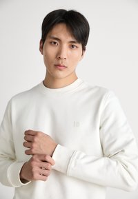 Λευκή φούτερ μπλούζα με ραμμένο λαιμό και μανσέτες, διαθέτει μικρή ετικέτα λογότυπου. Απαλή υφή υφάσματος με άνετη εφαρμογή.