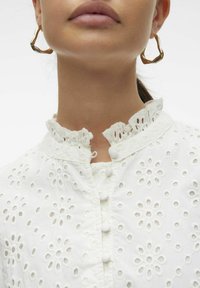 Blusa bianca ricamata con motivi floreali a intaglio, colletto arricciato e chiusura a bottoni. Il tessuto ha un aspetto texturizzato.