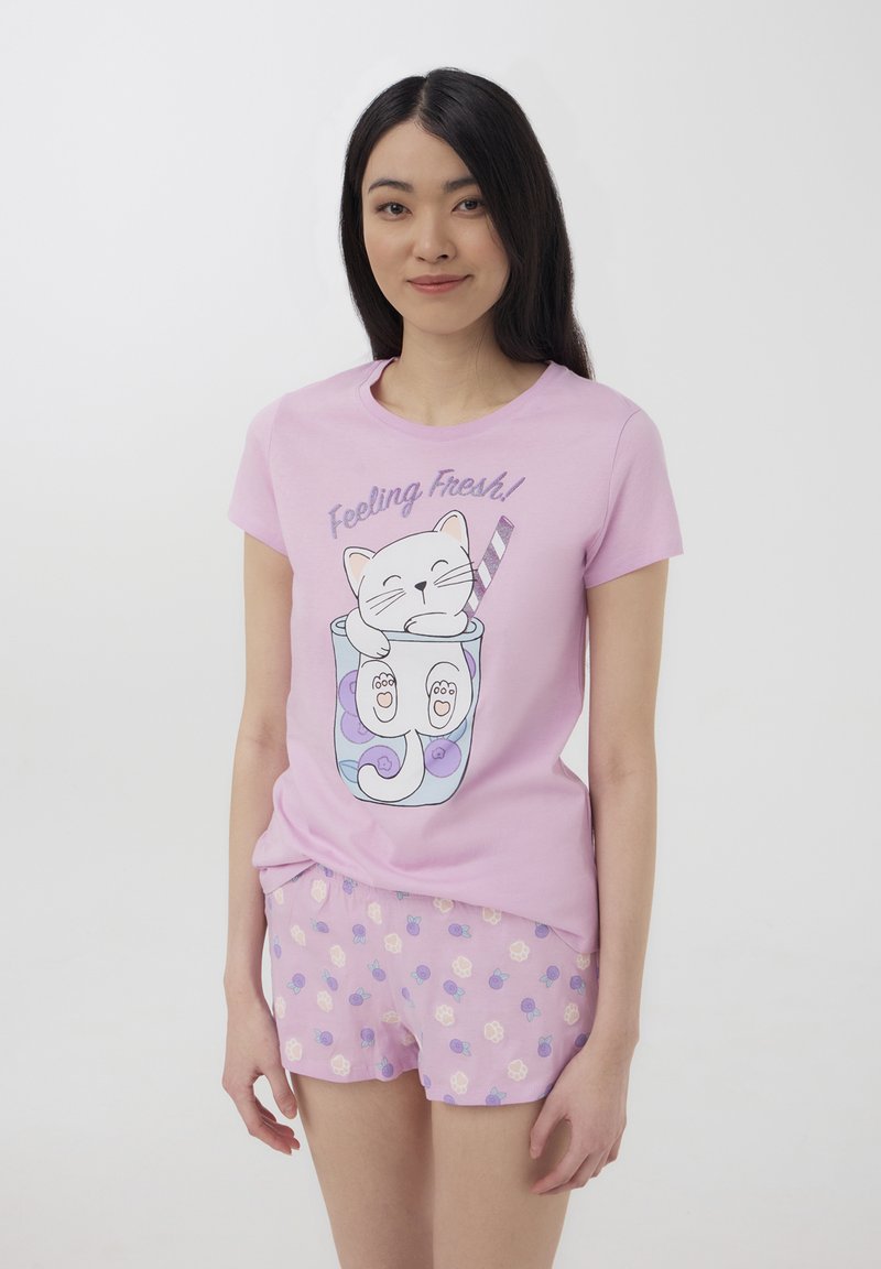 T-shirt di cotone rosa chiaro con un cartone animato di un gatto dentro una tazza con una cannuccia, abbinato a pantaloni corti rosa decorati con un motivo floreale in viola e crema.