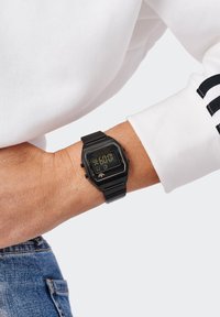 Montre numérique avec un boîtier et un bracelet rectangulaires noirs, affichant un écran numérique jaune. Portée au poignet au-dessus d'une chemise blanche à manches longues.