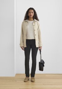 Chaqueta ligera beige con botones, suéter crema, pantalones negros ajustados y zapatos de lona color beige. Llevando un pequeño bolso negro.