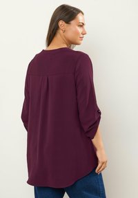 Bordeauxfarbendes Langarmhemd mit strukturiertem Design, abgerundetem Saum, Rückenfalte und hochgekrempelten Ärmeln. Kombiniert mit blauen Jeans.