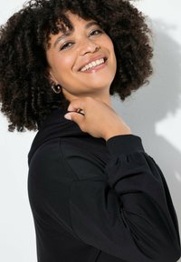 Černá mikina s měkkou texturou, s nastavitelnou kapucí a nařasenými manžetami. Model pózuje se ruka na rameni, což ukazuje uvolněný střih.