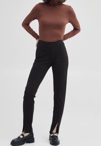 Un pantalon noir ajusté avec des fentes latérales au niveau des chevilles, associé à un haut à col roulé marron. La tenue est complétée par des mocassins noirs épais.