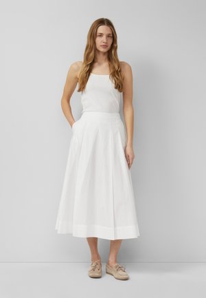 Kobieta z długimi włosami, ubrana w biały top bez rękawów i białą spódnicę midi, stojąca z jedną ręką w kieszeni i beżowymi butami.