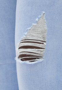 Ανοιχτό μπλε τζιν παντελόνι με μια μεγάλη κάθετη σχισμή που αποκαλύπτει σκούρο δέρμα από κάτω μέσα από ξεφτισμένες λευκές κλωστές.