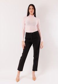 Maglione rosa chiaro a collo alto con texture a coste, abbinato a pantaloni neri sartoriali. La modella indossa tacchi trasparenti, con le mani posizionate ai lati.