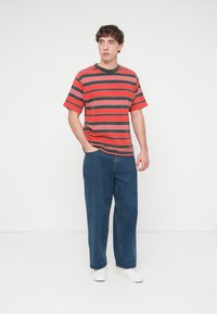 Κόκκινο και γκρι ριγέ μπλουζάκι με κοντά μανίκια, συνδυασμένο με φαρδιά μπλε τζιν και λευκά αθλητικά παπούτσια. Απλό φόντο.