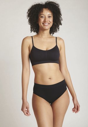 Lächelnde Frau mit lockigem Haar, die ein schwarzes Bralette und schwarze High-Waist-Unterwäsche trägt und vor einem einfarbigen Hintergrund steht.