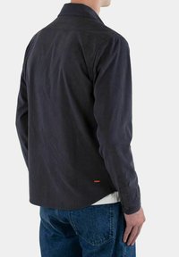Camicia in corduroy grigio scuro con colletto a punta, polsini con bottoni e un sottile logo arancione vicino all'orlo, che mette in mostra una superficie texturizzata.
