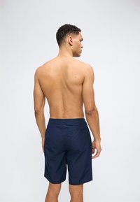 Άνδρας με κοντά σγουρά μαλλιά στέκεται με την πλάτη, χωρίς μπλούζα, φορώντας σκουρό μπλε μαγιό με πίσω τσέπη, σε απλό φωτεινό φόντο.