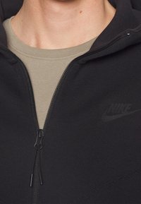 Svart zip-up hoodie med mjuk textur, förstärkta sömmar och en subtil ton-i-ton-logotyp på bröstet. Lager på en ljusbeige skjorta.