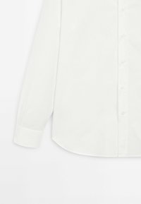 Chemise blanche à manches longues en tissu lisse, dotée d'un col classique, d'une patte de boutonnage devant et d'un ourlet arrondi. Les poignets sont repliés soigneusement.