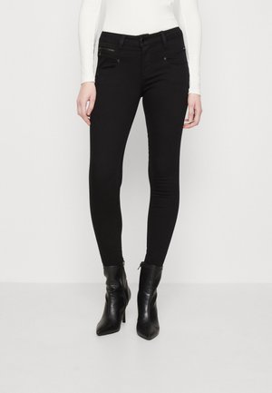Femme portant un jean skinny noir avec des poches zippées et des bottines noires à talons, debout sur un fond blanc uni.