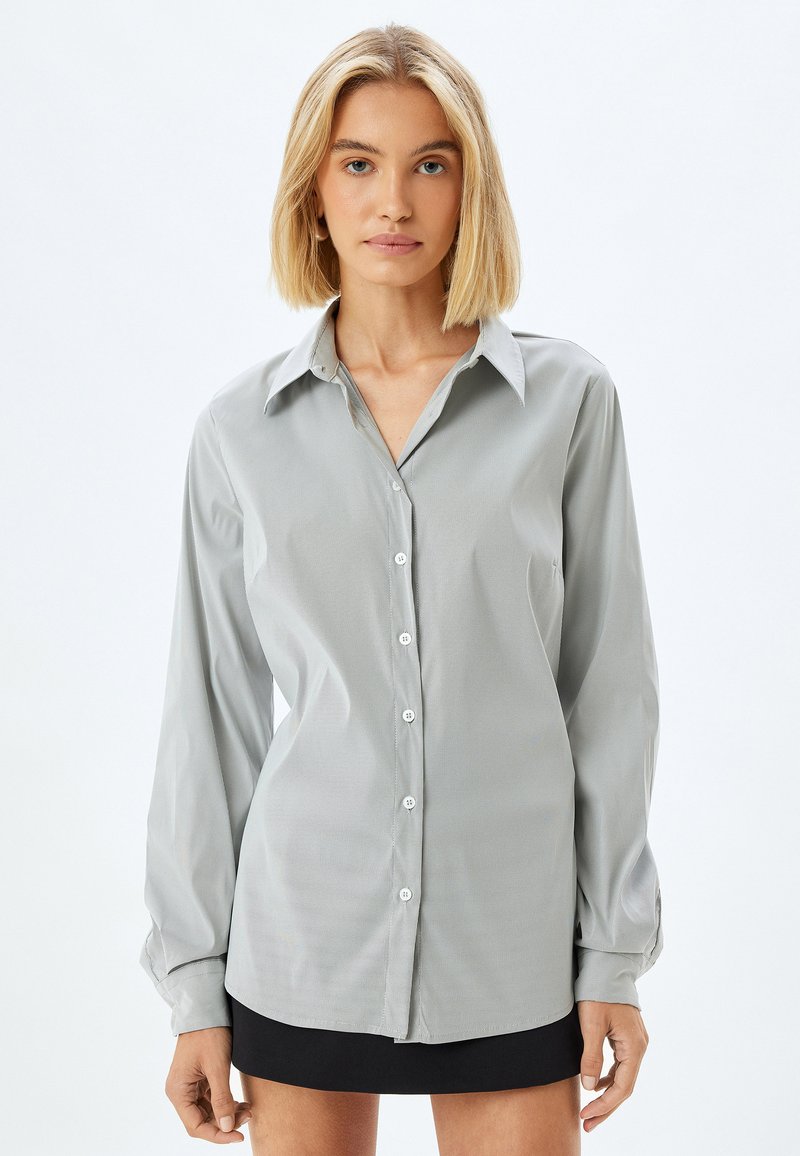 Camisa de botones de manga larga en gris claro, con cuello, que presenta un sutil patrón de rayas y un dobladillo redondeado, fabricada en una tela suave.