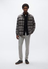 Γκρι πουκάμισο-μπουφάν με καρό σχέδιο και κουμπιά, μαύρο πουκάμισο από μέσα, ανοιχτό γκρι παντελόνι και σκούρα αθλητικά παπούτσια με καφέ σόλες.