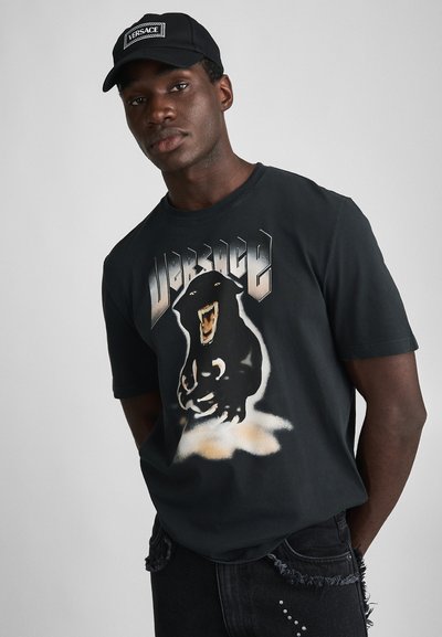Black t-shirt featuring a graphic of a roaring panther with the word "Versace" above. Styled with distressed black jeans and a black cap.