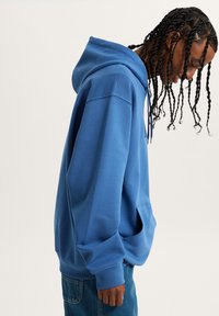 Sudadera de algodón azul con un corte relajado, gran bolsillo, capucha con cordón y puños acanalados. La tela tiene una textura suave y lisa.