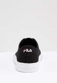 Sneaker nera FILA con suola bianca, logo visibile sul tallone, ripresa da dietro su una superficie bianca con sfondo grigio chiaro.