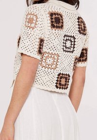 Žena v krátkém rukávu háčkovaném kardiganu s hnědými a béžovými čtvercovými vzory přes bílé sukni.