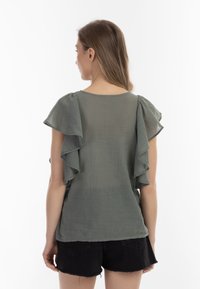 Oliven grøn, let bluse med lagdelte flæser på ærmerne, rund halsudskæring og subtilt gennemsigtig tekstur, parret med sorte shorts.