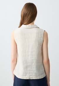 Top senza maniche in lino, colore beige chiaro, con colletto a punta e una texture sottile. Dettagli delle cuciture lungo l'orlo e sul retro.