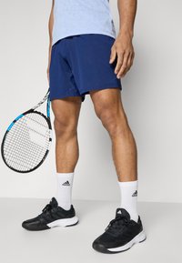 Shorts de sport bleu marine en tissu texturé, associés à des baskets noires et des chaussettes blanches de type crew avec un logo. Une raquette de tennis est tenue.