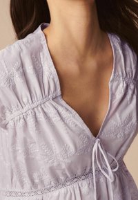 Robe en coton lilas avec broderie florale. Décolleté en V, corsage froncé et lien ajustable. Douces touches de dentelle le long de la taille.